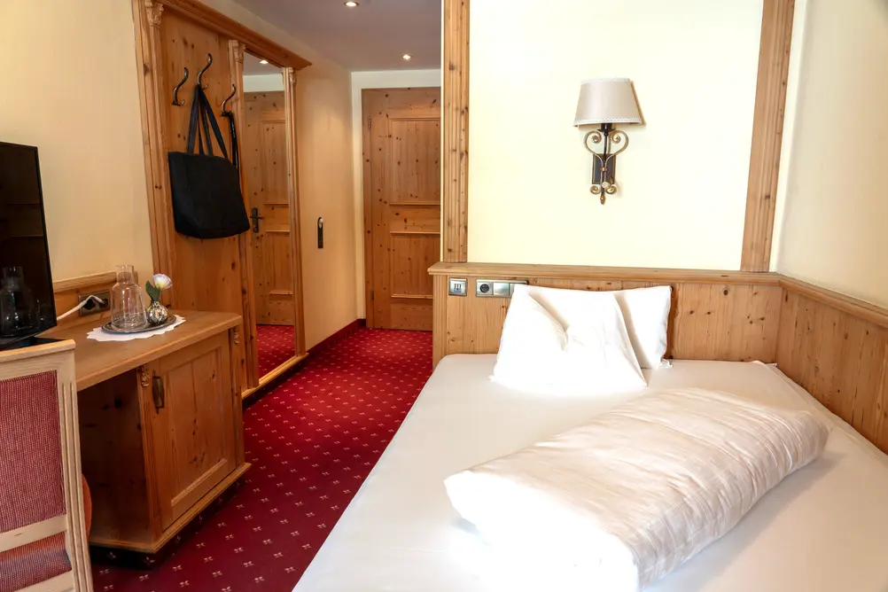 Traditionelles Hotelzimmer mit Einzelbett, Holzverkleidung, rotem Teppichboden, Kleiderschrank und Schreibtisch mit TV