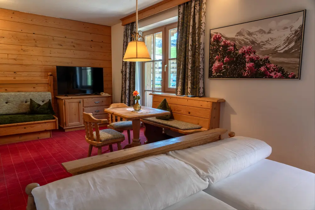 Alpenländisches Hotelzimmer mit Holzverkleidung, Sitzgruppe, TV, rotem Teppich und Bergblick-Balkon