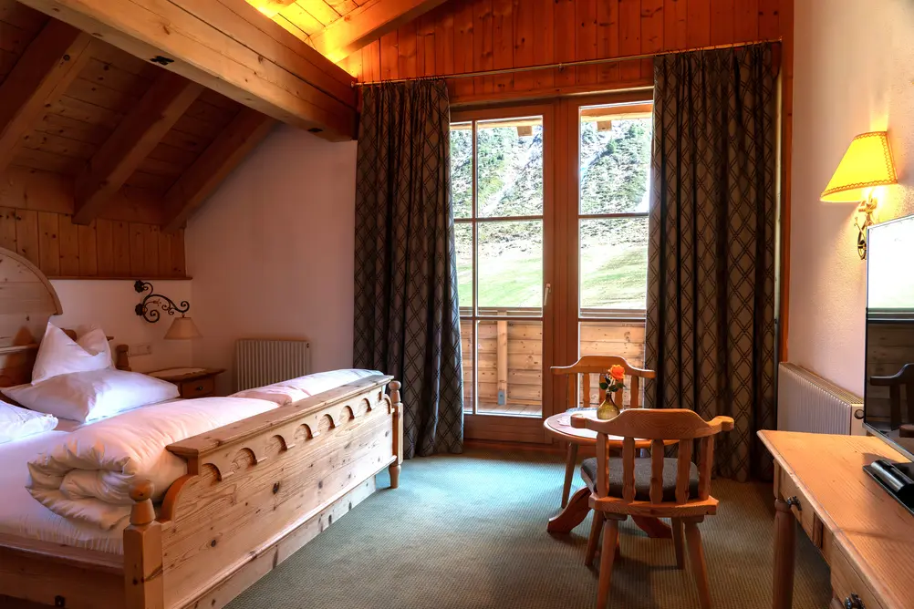 Alpines Hotelzimmer mit Holzdoppelbett, Sitzecke und Fenstertür zum Balkon mit Blick ins Grüne