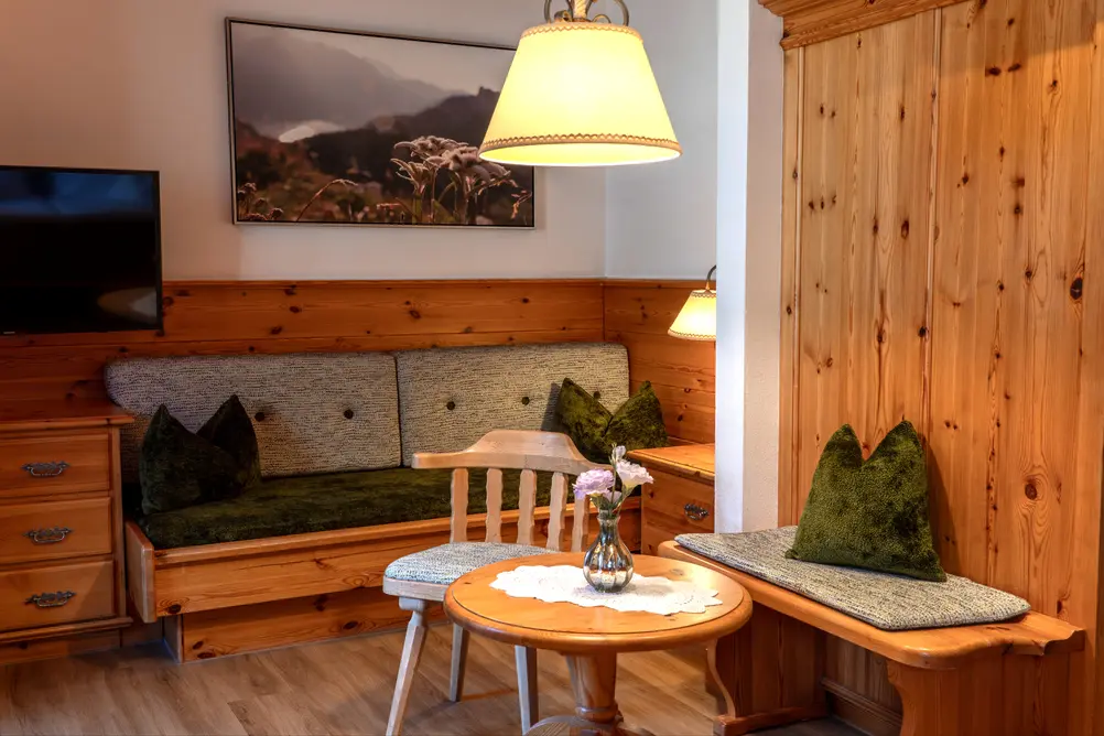 Eine rustikale Sitzecke mit einer eingebauten Holzbank, grünen Kissen, einem runden Holztisch mit einer Blumenvase, einem Wandbild mit Alpenblumen und einem hängenden Lampenschirm
