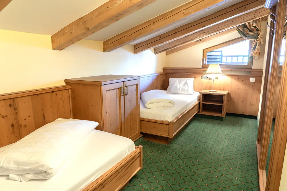 Ein gemütliches Dachgeschoss-Schlafzimmer mit zwei getrennten Holzbetten, Dachbalken, grünem Teppich und Holzverkleidung, beleuchtet von einer Stehlampe an einem kleinen Fenster