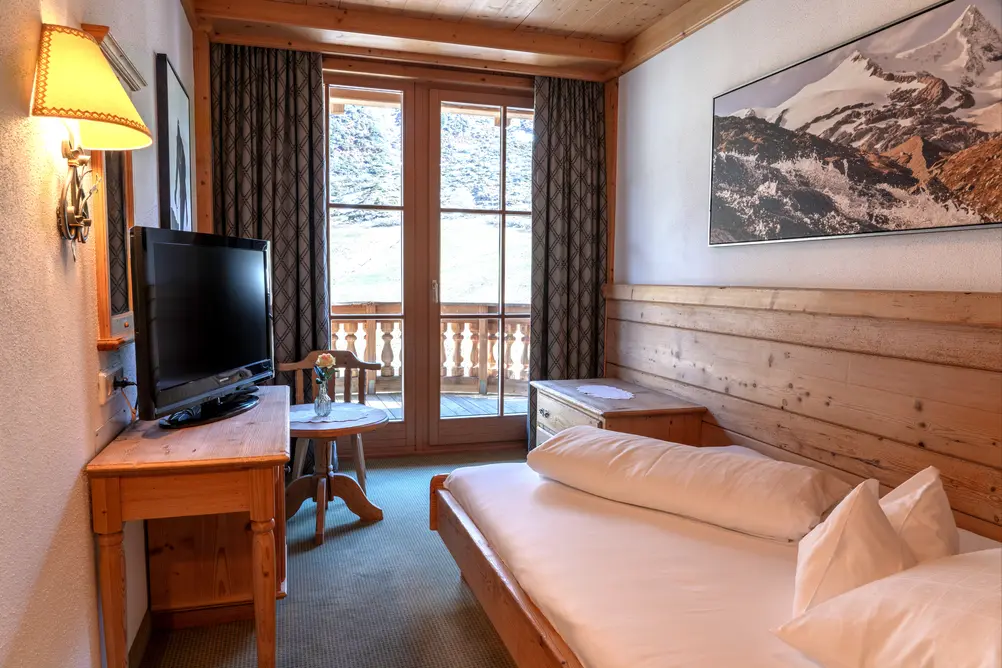 Holzmöbliertes Hotelzimmer mit Einzelbett, Fernseher, Tisch und Balkontür mit Bergblick