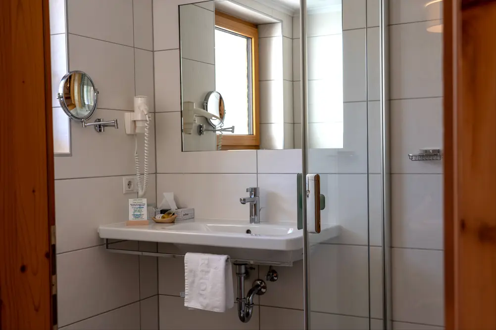 Hell gefliestes Hotelbadezimmer mit Waschbecken, Wandspiegel, Föhn und Glasdusche