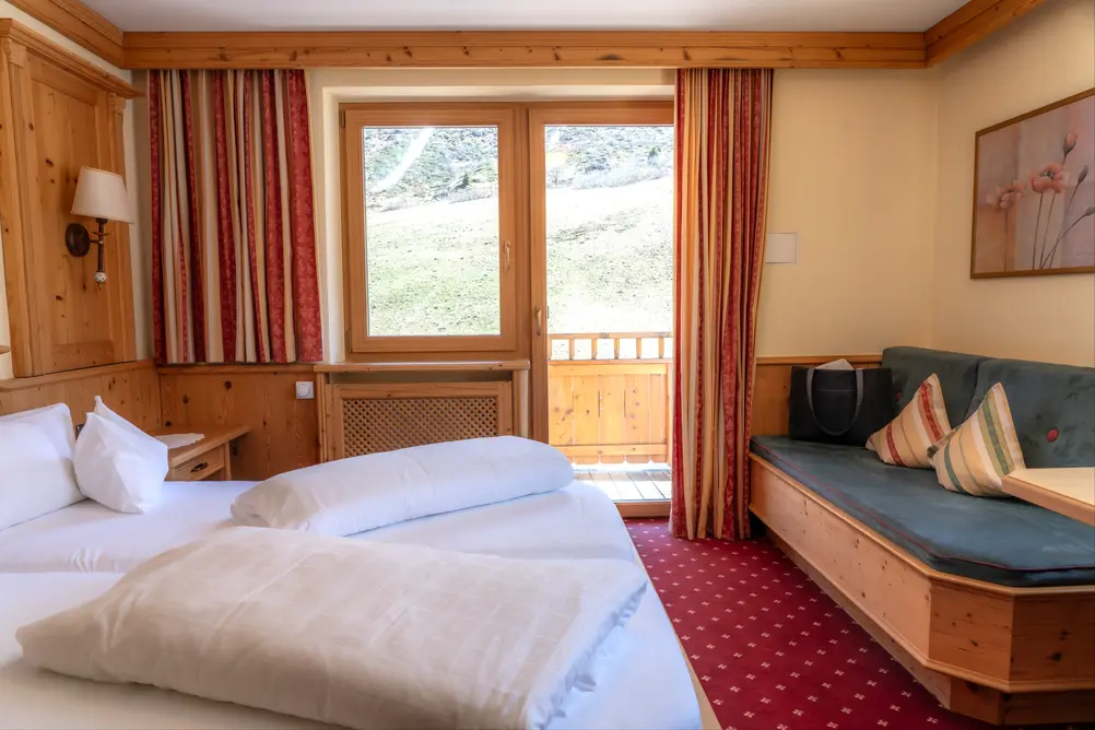 Gemütliches Hotelzimmer mit Holzmöbeln, Doppelbett, grünem Sofa und Balkontür ins Grüne