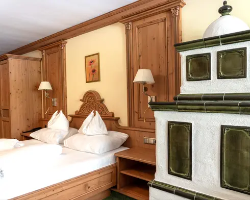 Doppelbett aus Holz mit weißer Bettwäsche neben einem traditionellen weiß-grünen Kachelofen