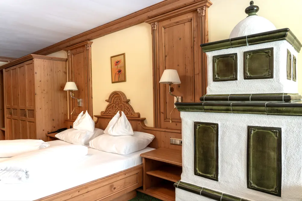 Doppelbett aus Holz mit weißer Bettwäsche neben einem traditionellen weiß-grünen Kachelofen