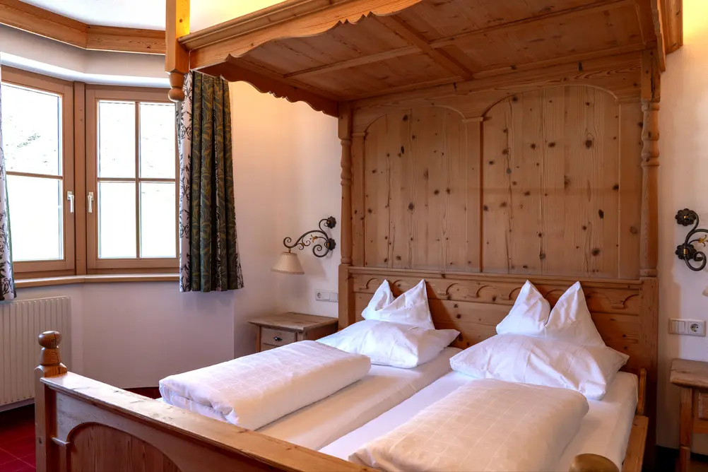 Rustikales Hotelzimmer mit großem Holz-Himmelbett, Nachttisch und Eckfenster