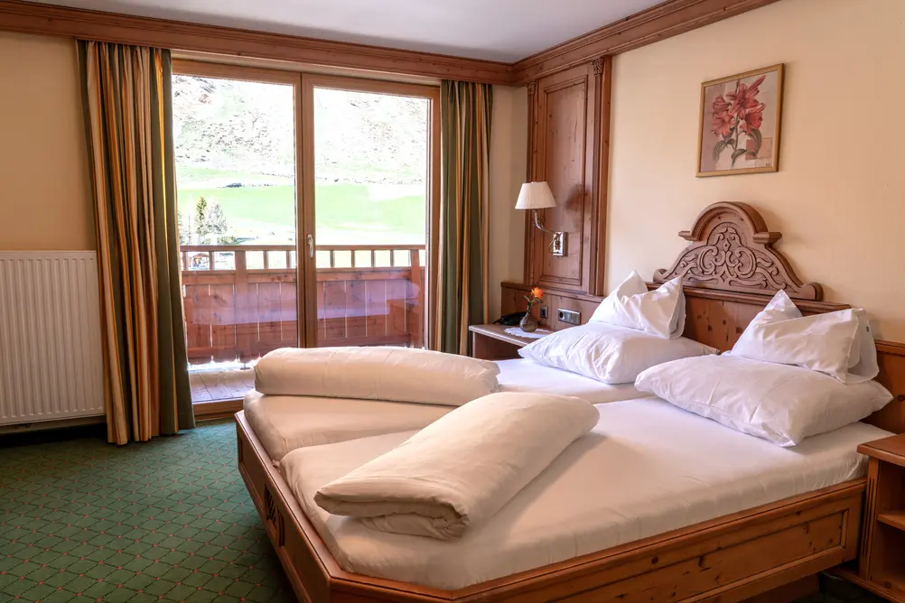 Ein helles Hotelzimmer mit einem geschnitzten Holz-Doppelbett, weißen Bettdecken, einem großen Glasschiebetür-Zugang zum Balkon mit Blick auf grüne Hügel, einem grünen Teppich und einem Blumenbild an der Wand