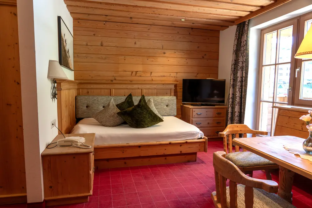 Blick in ein Zimmer mit Holzvertäfelung, bezogenem Bett, Fernseher und Sitzgruppe